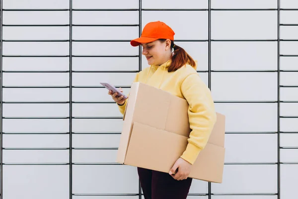 Young woman picks up parcel from automatic post office machine, Courier standing with phone and small box. Concept of fast delivery to automatic self lockers