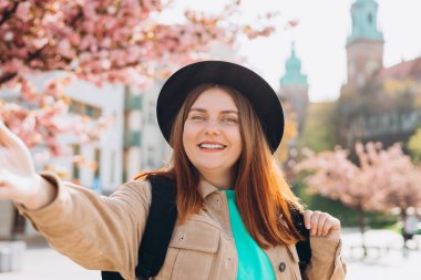 Neşeli, güzel bir kadın akıllı telefonuyla kameraya bakıyor açık havada portre fotoğrafı çekiyor. Krakow 'daki Wawel Kalesi yakınlarında siyah şapkalı kızıl saçlı bir kız. İlkbaharda Avrupa 'yı gezmek