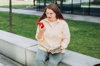 Üzücü, sarı tişörtlü, telefon kulübesinde oturan üzgün kız. Negatif insanların duyguları. Üzgün 30 'lu yaşlardaki genç bir kadın açık havada akıllı telefon kullanıyor.