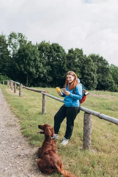 Happy redhead Woman in active trekking clothes having a halt after hiking. Hiker with backpack holding smart phone and drinking water from water bottle or hot drink from thermos