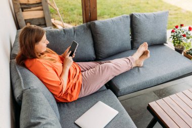 Neşeli, mutlu, genç, kızıl saçlı kadın yazın evin bahçesinde telefonla rahatlıyor. Sıradan, güzel bir kadın evdeki kanepede oturuyor.