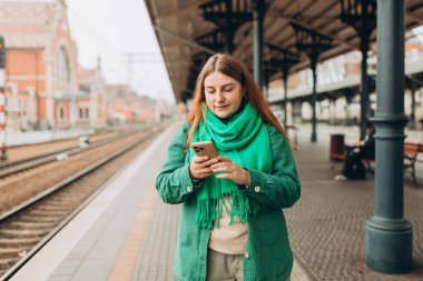 Sırt çantalı genç ve güzel bir kadın cep telefonu kullanırken tren bekliyor. Uygulama yoluyla cep telefonu izleme programını kullanan yolcu bayan
