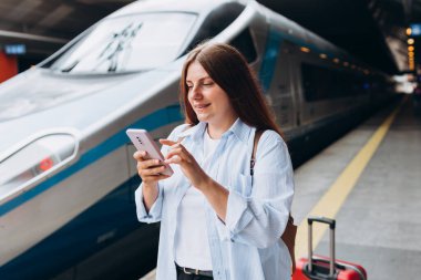 Sırt çantalı ve akıllı telefon kullanan genç, kızıl saçlı bir kadın. Demiryolu taşımacılığı konsepti, Gezgin. Tren istasyonunda bavullu kadınlar yürüyor. Tatile trenle git.