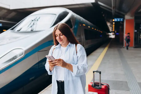 Young redhead woman waiting train with backpack and using smart phone. Railroad transport concept, Traveler. Women with suitcase walking at railroad station platform. Travel to vacation by train