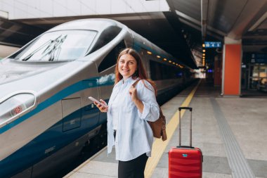 Sırt çantalı ve akıllı telefon kullanan genç, kızıl saçlı bir kadın. Demiryolu taşımacılığı konsepti, Gezgin. Tren istasyonunda bavullu kadınlar yürüyor. Tatile trenle git.