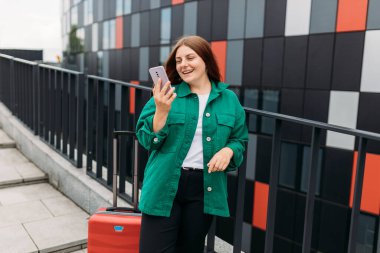 Young woman with a red suitcase using smart phone at airport. Redhead girl in international airport walking with her luggage.