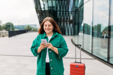 Young woman with a red suitcase using smart phone at airport. Redhead girl in international airport walking with her luggage.