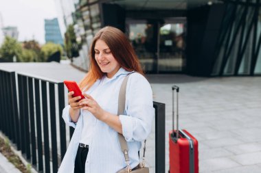 Young woman with a red suitcase using smart phone at airport. Redhead girl in international airport walking with her luggage.