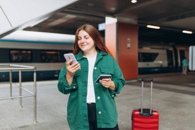 Sırt çantalı ve akıllı telefon kullanan genç, kızıl saçlı bir kadın. Demiryolu taşımacılığı konsepti, Gezgin. 30 'lu yaşlarda bavullu bir kadın tren istasyonunda yürüyor..