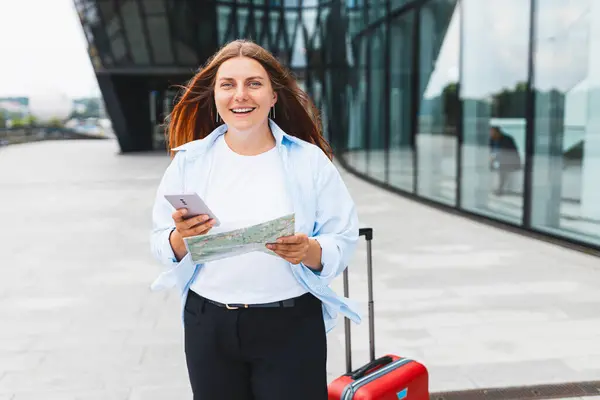 Beautiful redhead woman holding paper map and searching information on the internet with smartphone outdoors. Lady with suitcase choosing route via paper map