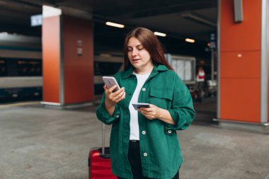 Sırt çantalı ve akıllı telefon kullanan genç, kızıl saçlı bir kadın. Demiryolu taşımacılığı konsepti, Gezgin. 30 'lu yaşlarda bavullu bir kadın tren istasyonunda yürüyor..