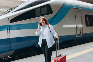 Sırt çantalı ve akıllı telefon kullanan kızıl saçlı kadın treni bekliyor. Demiryolu taşımacılığı konsepti. 30 'lu yaşlarda bavullu, akıllı telefondan konuşan, tren istasyonunda yürüyen orta yaşlı bir kadın.