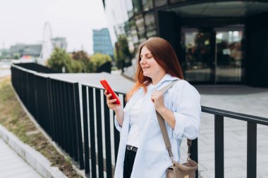 Genç mutlu iş kadını şehirde gülümsüyor ve akıllı telefon kullanıyor. Ofise giden güzel bir kadın akıllı telefonuyla internetten bilgi arıyor..