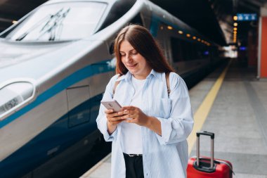 Sırt çantalı ve akıllı telefon kullanan genç, kızıl saçlı bir kadın. Demiryolu taşımacılığı konsepti, Gezgin. Tren istasyonunda bavullu kadınlar yürüyor. Tatile trenle git.