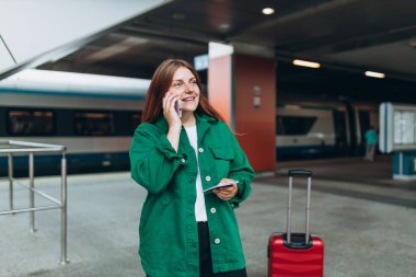 Kızıl saçlı kadın treni bekliyor ve akıllı telefon kullanıyor. Demiryolu taşımacılığı konsepti, Gezgin. 30 'lu yaşlarda bavullu, akıllı telefondan konuşan, tren istasyonunda yürüyen orta yaşlı bir kadın.