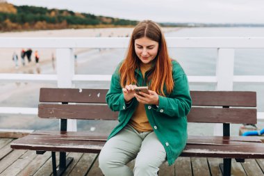 Dışarıda bankta oturan akıllı telefonlu, mutlu, 30 'lu yaşlarda genç bir turist. Sahilde yürüyen bir kadın. Yüksek kalite fotoğraf