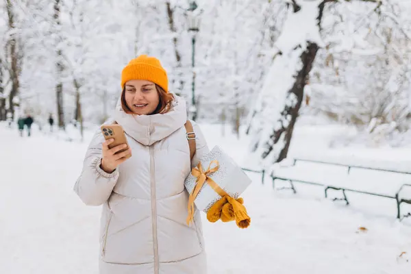 30 'lu yaşlarda, gülümseyen şık bir kadının portresi. Kışın şehir parkının dışında duruyor. Elinde telefon olan genç kadınlar Noel hediyelerini açık havada tutuyorlar.