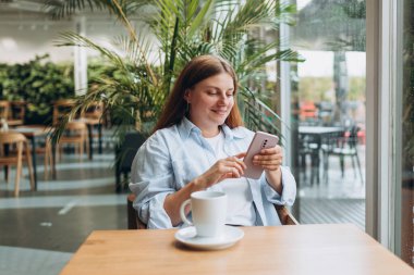 Genç mutlu iş kadını bir kafede gülümsüyor ve akıllı telefon kullanıyor. Kahve masasında oturan ve kapalı alanda akıllı telefon kullanan biri. Çevrimiçi eğitim, düzen, çalışma veya alışveriş kavramı