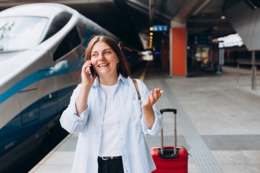 Kızıl saçlı kadın treni bekliyor ve akıllı telefon kullanıyor. Demiryolu taşımacılığı konsepti, Gezgin. 30 'lu yaşlarda bavullu, akıllı telefondan konuşan, tren istasyonunda yürüyen orta yaşlı bir kadın.