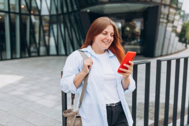 Genç mutlu iş kadını şehirde gülümsüyor ve akıllı telefon kullanıyor. Ofise giden güzel bir kadın akıllı telefonuyla internetten bilgi arıyor..