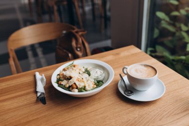 İçinde kızarmış ekmek, tavuk ve parmesan peyniri olan beyaz bir kase dolusu Cezar salatası. Tahta masanın üzerinde beyaz tabakta Cezar salatası