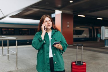 Kızıl saçlı kadın treni bekliyor ve akıllı telefon kullanıyor. Demiryolu taşımacılığı konsepti, Gezgin. 30 'lu yaşlarda bavullu, akıllı telefondan konuşan, tren istasyonunda yürüyen orta yaşlı bir kadın.