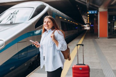 Sırt çantalı ve akıllı telefon kullanan genç, kızıl saçlı bir kadın. Demiryolu taşımacılığı konsepti, Gezgin. Tren istasyonunda bavullu kadınlar yürüyor. Tatile trenle git.