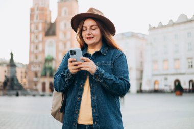 Çekici genç bayan turist yeni şehri keşfediyor. Krakow 'daki Market Meydanı' nda elinde akıllı bir telefon tutan kızıl saçlı kız. Yazın Avrupa 'yı geziyorum. Aziz Mary Bazilikası.