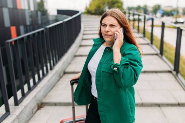 Kırmızı bavullu, mutsuz, kızıl saçlı genç iş kadını şehirde cep telefonuyla arama yapıyor. Cep telefonuna bağıran öfkeli bir kadının portresi.