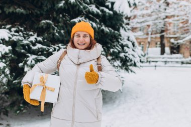 Mutlu kadın baş parmağını kaldırır, kışın şehir parkında hediye kutusunu tutar. 30 'lu yaşlardaki genç kadınlar Noel hediyelerini açık havada tutuyorlar. Alışveriş kavramı, tatiller