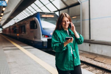Kızıl saçlı kadın treni bekliyor ve akıllı telefon kullanıyor. Demiryolu taşımacılığı konsepti, Gezgin. 30 'lu yaşlarda orta yaşlı bir kadın akıllı telefondan konuşuyor, tren istasyonunda yürüyor.