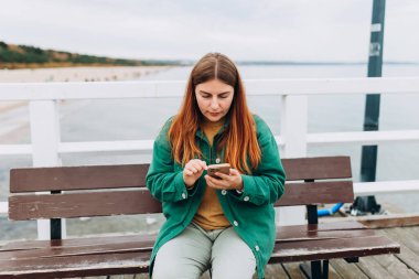 Dışarıda bankta oturan akıllı telefonlu, mutlu, 30 'lu yaşlarda genç bir turist. Kızıl saçlı kadın rıhtımda yürüyor, deniz kenarında.