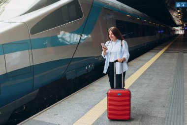 Sırt çantalı ve akıllı telefon kullanan genç, kızıl saçlı bir kadın. Demiryolu taşımacılığı konsepti, Gezgin. Tren istasyonunda bavullu kadınlar yürüyor. Tatile trenle git.