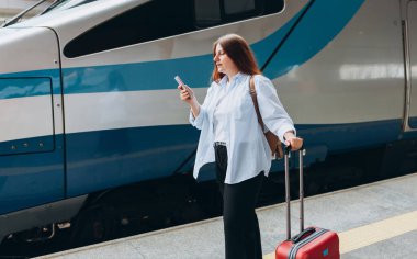 Sırt çantalı ve akıllı telefon kullanan genç, kızıl saçlı bir kadın. Demiryolu taşımacılığı konsepti, Gezgin. Tren istasyonunda bavullu kadınlar yürüyor. Tatile trenle git.