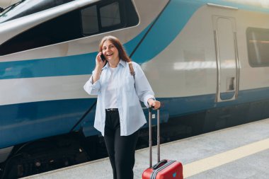 Sırt çantalı ve akıllı telefon kullanan kızıl saçlı kadın treni bekliyor. Demiryolu taşımacılığı konsepti. 30 'lu yaşlarda bavullu, akıllı telefondan konuşan, tren istasyonunda yürüyen orta yaşlı bir kadın.