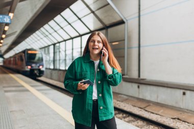 Kızıl saçlı kadın treni bekliyor ve akıllı telefon kullanıyor. Demiryolu taşımacılığı konsepti, Gezgin. 30 'lu yaşlarda orta yaşlı bir kadın akıllı telefondan konuşuyor, tren istasyonunda yürüyor.