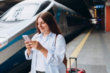 Sırt çantalı ve akıllı telefon kullanan genç, kızıl saçlı bir kadın. Demiryolu taşımacılığı konsepti, Gezgin. Tren istasyonunda bavullu kadınlar yürüyor. Tatile trenle git.