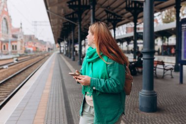 Sırt çantalı ve akıllı telefon kullanan genç, kızıl saçlı bir kadın. Demiryolu taşımacılığı konsepti, Gezgin. 30 'lu yaşlardaki kadın tren istasyonunda yürüyor.