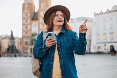 Çekici genç bayan turist yeni şehri keşfediyor. Kızıl saçlı kız Krakow 'daki Market Meydanı' nda bir akıllı telefon tutuyor. Yazın seyahat. Aziz Mary Bazilikası.