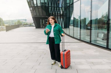 Young woman with a red suitcase using smart phone at airport. Redhead girl in international airport walking with her luggage.