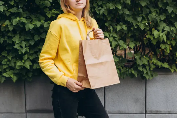 Girl with bag in hand posing on city background. A Female delivery hand with craft shopping bag outdoors. Fast Food delivery service, area for your logo or design. Packaging mock up.