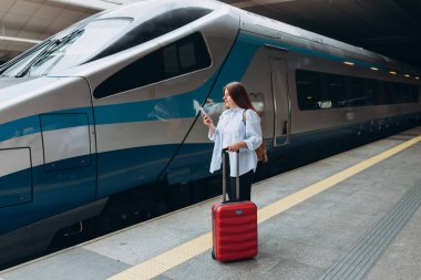 Sırt çantalı ve akıllı telefon kullanan genç, kızıl saçlı bir kadın. Demiryolu taşımacılığı konsepti, Gezgin. Tren istasyonunda bavullu kadınlar yürüyor. Tatile trenle git.