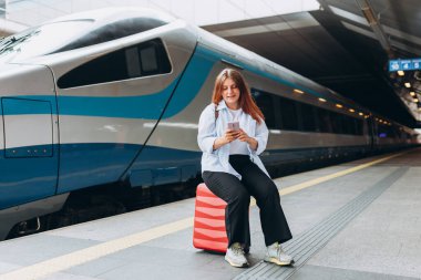 Sırt çantalı ve akıllı telefon kullanan genç, kızıl saçlı bir kadın. Demiryolu taşımacılığı konsepti, Gezgin. Tren istasyonunda bavullu kadınlar yürüyor. Tatile trenle git.