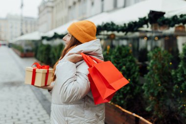 Woman holding red shopping paper bags and present in hands, walking at street. Winter holidays sale. Shopping banner, for mockup