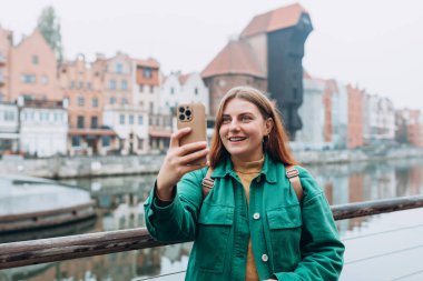 Happy 30s Women taking selfie on urban background. Young beautiful girl say Hi. Gdansk old town and famous Zuraw crane, A beautiful woman is standing near Motlawa river clipart