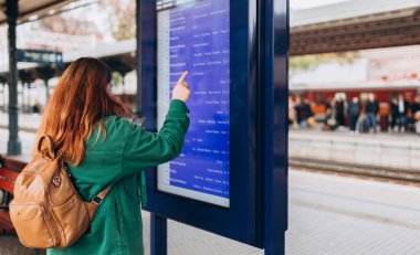 Sırt çantalı genç kızıl saçlı kadın tren istasyonunda tren tarifesini işaret ediyor. 30 'lu yaşlardaki kadın Geliş ve Gidiş Bilgileri Görünümüne bakıyor