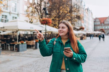 Şehir caddesinde telefonuyla yürüyen mutlu, genç bir kadın akıllı telefonunu kontrol ediyor. Sırt çantalı kızıl saçlı kız şehir caddesini işaret ediyor. Avrupa 'yı gezmek