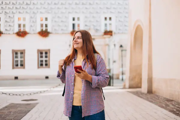 Picture of pretty young woman staying on the street holding phone in hands. 30s tourist walking on old city street checks her smartphone. Use technology concept