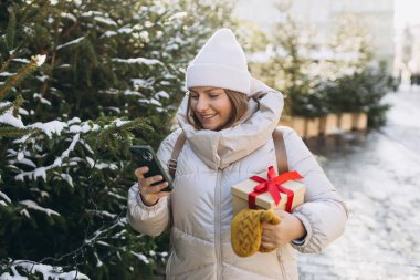 Kış aylarında şehir parkında 30 'lu yaşlarda, gülümseyen şık bir kadın. Akıllı telefonları olan genç kadınlar Noel hediyelerini açık havada tutuyorlar. Telefon iletişimi. İnternette gezinen kız