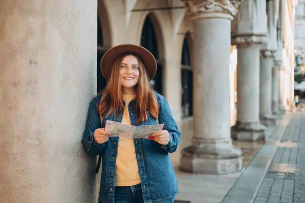 Pretty young woman staying on the street holding paper map in hands. 30s girl in hat posing on Market Square in Krakow. High quality photo, The Cloth Hall, Vacation concept